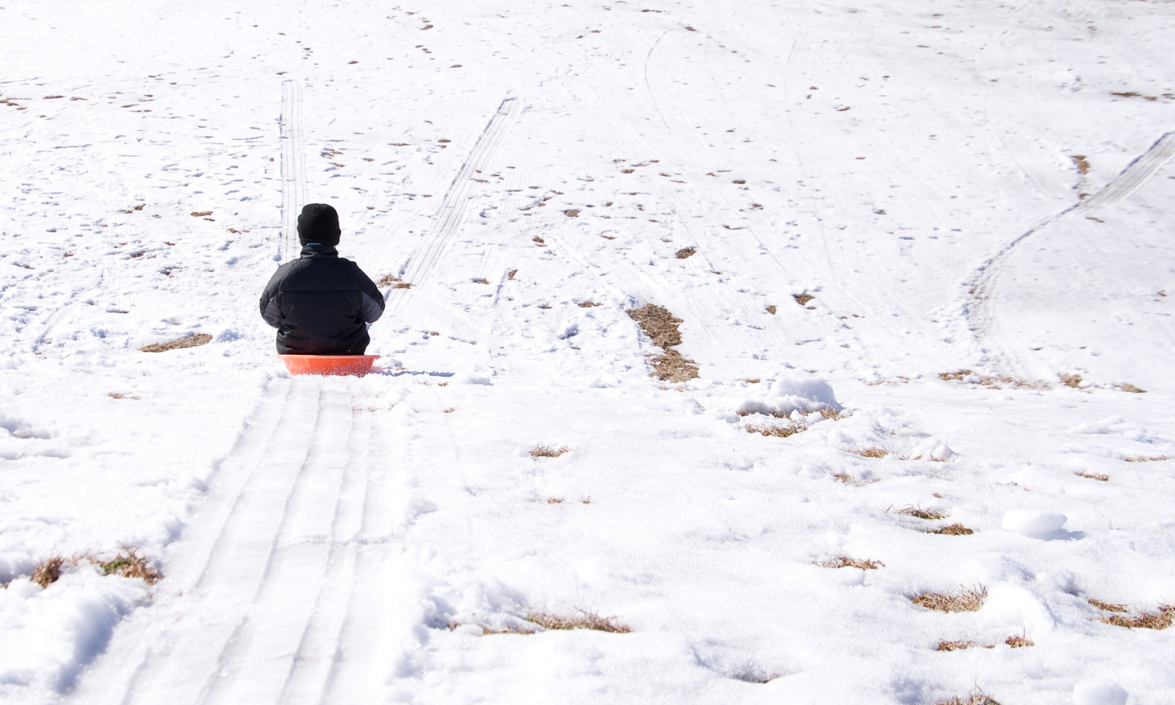 Rear-view image of a child sledding down a snowy hill with a black coat